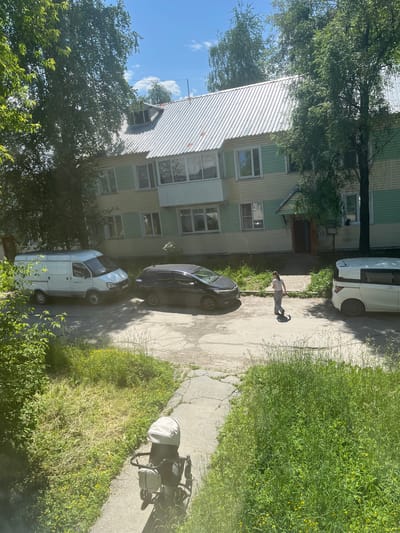 The image depicts a residential building in Baykalsk, Russia, on a sunny day. The building is a multi-story structure with a light-colored, possibly metal, corrugated roof. The facade is painted in alternating horizontal bands of light yellow and pale green. There are several windows and a balcony visible.
In front of the building, on a paved area that appears to be a courtyard or parking lot, are several vehicles. To the left, a white cargo van is parked. Next to it, to the right, is a dark-colored sedan. Further to the right, a white minivan is parked.
A young person, possibly a girl, is walking in the middle of the paved area, casting a shadow. She is wearing a light-colored t-shirt and dark pants.
The scene is surrounded by lush green trees and vegetation, with grass and some small yellow flowers growing along the edges of the paved area and on a grassy embankment. A baby stroller is positioned on the paved pathway in the foreground, facing away from the camera. The sky above is blue with scattered white clouds.