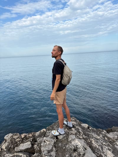 A medium-shot photograph shows a man standing on a rocky cliff overlooking the ocean. The sky is visible in the upper left of the photo, featuring clouds, with a line defining the horizon visible in the center. The man stands near the edge of the cliff, wearing a black t-shirt, beige shorts, white socks, and patterned clogs. He has a light grey backpack on his back. He is looking down at the ocean. The cliff is made of jagged gray rocks. The location is Bondarenkovo, Ukraine.