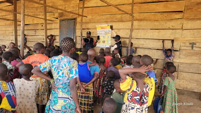 In the city of Bunia, Democratic Republic of the Congo, a group of children are gathered in a makeshift wooden structure for an awareness session. The structure has a wooden plank wall and a wooden beam ceiling, with a blue door visible on the left. Two men are addressing the children; one, wearing a red vest and a blue cap, holds a megaphone, while the other, in a blue vest and black cap, points to a poster on the wall. The poster, which appears to be about Ebola, has text and images. The children, varying in age and dressed in colorful clothing, are standing and looking towards the speakers. Some children are in the foreground, facing away from the camera, while others are in the background, some reaching out to touch the wooden walls. A few children are positioned on the left side, near the door, and some are on the right, near a child standing on a barrel. The overall scene depicts an educational outreach event in a community setting.