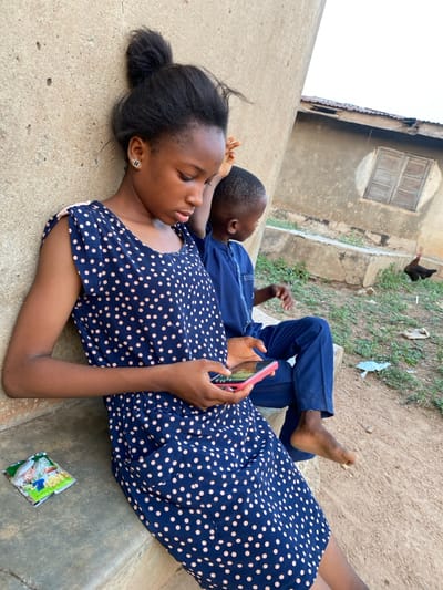 The image depicts two individuals, a young female and a young male, seated outdoors in Idiroko, Nigeria. The female, a teenager with dark skin and dark hair tied in a top bun, is positioned in the foreground and left. She wears a navy blue sleeveless dress with light pink polka dots and gold-colored clover stud earrings. She holds a pink-cased smartphone with both hands, her gaze directed downwards at the screen. To her right and slightly behind her, a younger boy with dark skin and short dark hair sits barefoot. He wears a navy blue traditional long-sleeved top and matching trousers, looking towards the right. Both are seated on a light gray concrete ledge that abuts a textured, light brown wall. A discarded green and yellow wrapper lies on the concrete ledge to the female's left. The background features an uneven dirt ground with sparse green vegetation and litter. Further back, a light brown building with a weathered, dark wooden-slatted window set within a circular frame is visible, along with part of a corrugated metal roof. A dark-feathered chicken stands on the ground near the building.