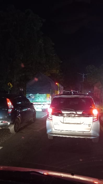 The image depicts a nocturnal traffic scene on a road in Tebel, Indonesia. The perspective is from inside a vehicle, looking forward through its windshield, with the top edge of a red-brown dashboard visible at the bottom of the frame.

The road is occupied by multiple vehicles experiencing congestion. Directly in front of the camera is a silver minivan, its rear illuminated by active brake or taillights. It features rear parking sensors and a partially visible Indonesian license plate starting with 'B'. To the immediate left of this minivan is a dark-colored hatchback or small SUV, its left rear taillight also illuminated.

Further ahead, a large truck with a dark tarpaulin-covered cargo bed is positioned in the same lane. Its rear displays a prominent light-colored advertisement or banner with text, including "UNTUK INDONESIA" and "KEBANGGAAN," suggesting a national pride theme. Beyond this truck, a line of additional vehicles, distinguishable by their illuminated lights, extends into the darkness, indicating further traffic.

The environment is heavily shadowed by night. Dark, dense foliage, consistent with trees and bushes, lines both sides of the road. On the left, a yellow diamond-shaped road sign with a black symbol (likely a warning or informational sign) is affixed to a pole amidst the trees. No individuals are clearly visible. The road surface appears dark, possibly asphalt, with minor reflections from vehicle lights.