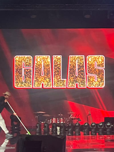 The image displays the word "CULTO" vertically stacked with each letter outlined in red and filled with a gold speckled pattern. A drum set and stage lights are visible on the right side, along with a stagehand holding a mop, possibly cleaning the floor. A red background subtly illuminates the scene.