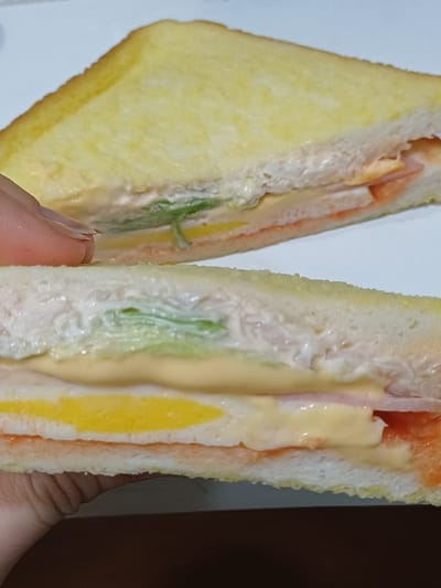 The scene, located in 서대문구, South Korea, depicts two halves of a sandwich, cut diagonally. A light-skinned human hand, with visible fingernails, holds the lower sandwich half, presenting it towards the viewer. The sandwich is constructed with two slices of light brown, possibly toasted, white bread. Visible internal layers, from top to bottom in the lower half, include a substantial white creamy spread, a green leafy vegetable, a thick yellow layer (presumed to be an egg patty or cheese slice), a thin slice of pink processed meat (likely ham), and a creamy yellow sauce. The upper sandwich half, partially obscured by the hand, shows similar layered fillings. The background is a plain white surface, suggesting an indoor setting, possibly a kitchen or dining area.