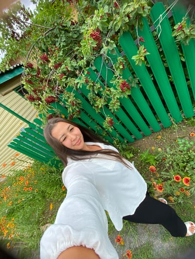 In Akyar, Russia, a young woman with long, dark hair is captured in a high-angle selfie, standing in a yard with a vibrant green wooden fence. She is wearing a white top and black leggings, and her right eye is winked. The fence is partially obscured by lush green branches laden with clusters of small, dark red berries. Beneath the branches and in front of the fence, a bed of small, bright orange and yellow flowers blooms. The ground is covered in a mix of grass and soil. To the left of the scene, a portion of a building with a light-colored, corrugated metal exterior and a green trim is visible. The overall setting suggests an outdoor, possibly residential, environment.