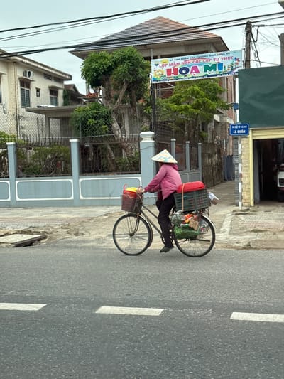 In Nam Đông Hà Ward, Vietnam, a person wearing a conical hat and a pink jacket rides a bicycle loaded with goods. The bicycle has two baskets attached, one in the front and one in the back. The rear basket is filled with green bags and other items, topped by a red woven container. The person is dressed in dark pants and green shoes.
The scene unfolds on a paved road with white lane markings. To the left of the road, a light blue wall with white decorative elements runs along a property, bordered by a metal fence. Behind the fence, a building with multiple windows and an air conditioning unit is visible, along with a large green tree.
In the background, a building with a dark-tiled roof is under construction or renovation, with scaffolding visible. A banner advertising "HOA MI" preschool hangs from the building. A blue street sign indicates "KIỆT 520 LÊ DUẨN". To the right, a building with a yellow facade and a garage-like opening is present. The sky is overcast.