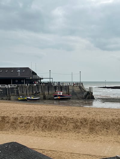This image depicts Ramsgate Harbour in Thanet, United Kingdom, at low tide under an overcast sky. The foreground consists of a broad, light brown sand beach exhibiting ripples and texture. In the midground, a substantial concrete and timber harbour wall extends from left to right, dividing the beach from the receding sea. Three boats are beached on the exposed wet sand and mudflats at the base of this wall. To the left, two smaller fishing vessels, one yellow and one blue-hulled with "F 497" discernible, rest on the exposed seabed. To the right, a larger blue-hulled boat with a red cabin and white trim is beached, with a lone individual wearing an orange lifevest standing beside its starboard side, facing the vessel.
The harbour wall features dark grey concrete sections with vertical wooden pilings, equipped with multiple wooden ladders and several rubber tires mounted for fendering. A section of concrete and wooden steps descends from the wall towards the water on the right. A walkway with white railings runs along the top of the wall, illuminated by black lampposts connected by string lights. A large, dark-roofed building, possibly a boathouse or maritime structure, stands on the harbour to the left. This building has multiple dormer windows on its roof and circular porthole-style windows on its dark walls, complemented by white exterior railings. In the background, the sea stretches to a low horizon under a predominantly grey, cloudy sky. Choppy water is visible, and a navigational marker, a pole with a flag-like element, is present in the distant water to the right. Indistinct figures are visible on the elevated walkway atop the harbour wall.