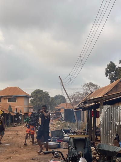 An overcast day in what appears to be a town or village. Multiple power lines stretch overhead, connected to a leaning utility pole. Several buildings, varying in height and construction, line the scene. A group of people are standing on a dirt ground, one shirtless, near what seems to be a motorcycle. In the background, other buildings and trees fill the horizon.