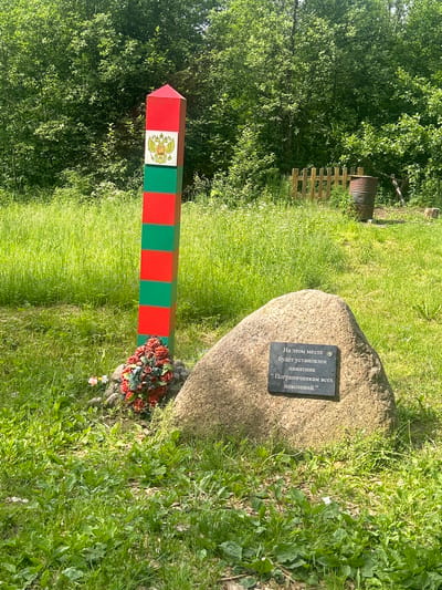 The image shows a border post and a memorial stone in a grassy area with trees in the background. The border post is red and green striped with the Russian coat of arms at the top. A wreath of red flowers is placed at the base of the post. The memorial stone is a large, rough-hewn rock with a plaque attached. The plaque is inscribed with Russian text. Behind the border post and stone, there is a wooden fence and a metal barrel. The scene appears to be a memorial site, likely related to border control. The inscription on the stone translates to: "On this spot a monument will be erected 'To the border guards of all generations.'"