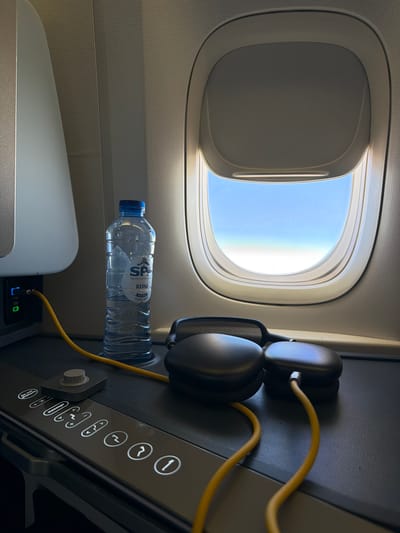 The image shows a close-up view of an airplane seat with a window. On the tray table, there is a bottle of SPA Reine mineral water, a pair of black headphones with a yellow cord, and a charging port with a cable plugged in. The tray table also features several illuminated buttons, including "Do not disturb," "massage," and numbered icons. The window reveals a clear blue sky. The scene is well-lit by natural light coming from the window. There are no people visible in the image. The location is identified as Rachel, United States, which is likely referring to the location of the photograph, not necessarily the subject matter itself.
