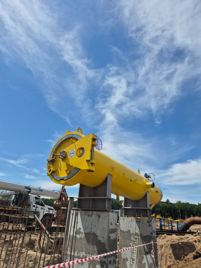 This is a full-shot, ground-level, exterior photograph taken on a sunny day with a blue sky and scattered white clouds. The scene appears to be a construction site in Taezhnoe, Russia, focusing on a large, bright yellow cylindrical vessel, possibly a gas separator, elevated on concrete and steel supports. The vessel is positioned diagonally, with its open end facing the left of the frame and its closed end to the right. It features several valves and a circular flange with bolts on its left end. A crane truck with the branding "STROMPROM" is visible to the left, partially obscured by construction materials and rebar. In the background, a line of blue and yellow pipes is installed, and a section of a large, dark-colored pipe bends into the foreground from the right. The ground is uneven and appears to be disturbed earth, with some concrete structures and rebar present in the foreground, indicating ongoing construction. The overall environment suggests an industrial infrastructure project. There are no people clearly visible in the image.