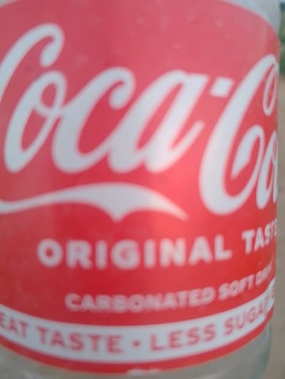 The image shows a close-up of a red Coca-Cola bottle, with the iconic Coca-Cola logo prominently displayed in white. Below the logo, text reads "ORIGINAL TASTE" and "CARBONATED SOFT DRINK". Further down, another line of text is partially visible, reading "GREAT TASTE - LESS SUGAR". The bottle appears to be made of clear glass. The background is blurred and out of focus, making it difficult to discern specific details of the environment. However, it appears to be an outdoor setting with some indistinct natural elements. The image is undated and the exact location in Rahwol Chom, Nigeria, cannot be determined from the visual information provided.