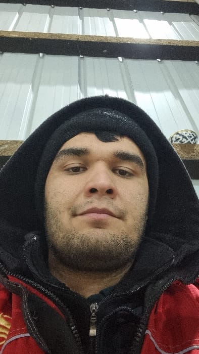 A close-up, chest-up image taken in Kagan, Uzbekistan, features an adult male facing the camera directly. The individual has dark hair visible beneath a black knitted beanie, dark eyes, and a short beard. He is dressed in a dark hooded garment over the beanie, with a partially zipped black fleece jacket visible beneath the hood. A red outer jacket, featuring yellow embroidery on the left side and white trim, is worn underneath the black fleece. The background above the subject reveals light-colored vertical corrugated metal panels, traversed by horizontal wooden beams. A small, round decorative object with a black and white geometric pattern rests on a shelf to the subject's upper right. The scene is illuminated by what appears to be artificial light.