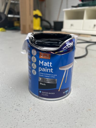 A partially opened can of black matte paint sits on a speckled gray concrete floor. The can's lid is bent and peeled back, revealing the thick black paint inside. The label on the can is blue and features text indicating "2IN1 Matt paint," "primer & finish in one," "wood / metal / PVC," "EXCELLENT PROTECTION," and the color "Black." It also shows icons for temperature and drying times, as well as "WATER BASED," "Low odour & quick dry," and "10 m²/l." In the blurred background, there is a yellow cleaning machine, a cardboard box, a white cabinet, and electrical cords. The scene appears to be indoors, possibly a garage or workshop. There are no people visible in the image. The lighting suggests it is daytime, with artificial light likely supplementing natural light.