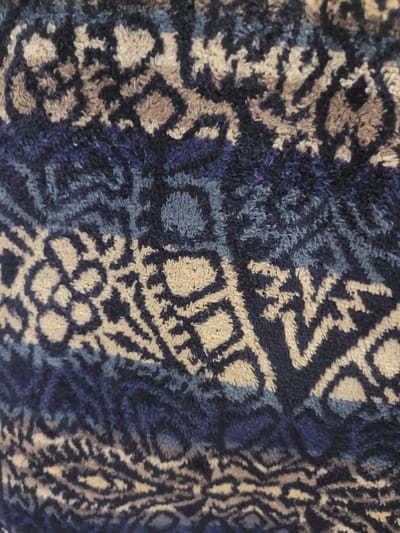 The image shows a close-up view of a textured fabric with a repeating abstract pattern in shades of dark blue, light blue, beige, and brown. The pattern consists of irregular shapes and lines that resemble organic forms or stylized animal prints. The fabric appears to be thick and possibly plush, such as a carpet or upholstery. There is no indication of any specific location in Gävle, Sweden, or any people, actions, or interactions.