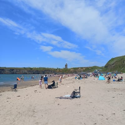 The image depicts Ballybunnion Beach in Ballybunnion, Ireland, on a bright, sunny day with a clear blue sky dotted with white clouds. A large number of people are enjoying various beach activities. In the foreground, the sandy beach is occupied by several beach chairs, towels, and a person sitting in a chair. Further back, people are walking, playing in the sand, and wading in the ocean. The water appears calm, with some individuals swimming or playing in the shallows. The middle ground shows a more densely populated area of the beach with numerous people sunbathing, picnicking, and congregating. Some umbrellas and tents are set up to provide shade. The background features a dramatic cliff face with vegetation, and on top of the cliff, a historic stone tower, likely a remnant of a castle, stands as a prominent landmark. The overall scene is one of a bustling, popular beach on a pleasant summer day.