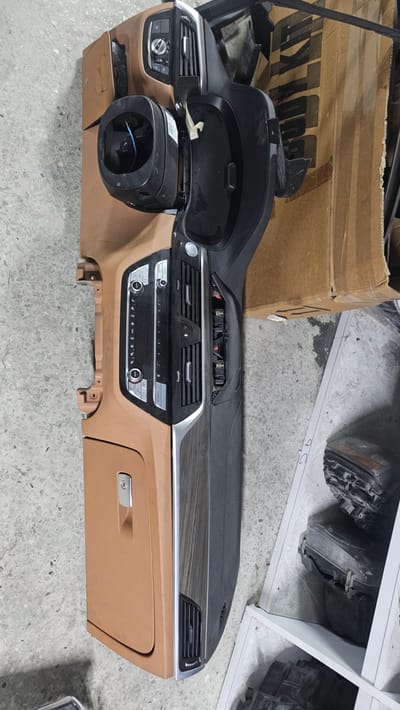 The media file shows a disassembled car dashboard, likely for a BMW, with a brown interior and black trim. The dashboard is lying on a concrete floor, with a large cardboard box in the background. Shelving units filled with car parts, including headlights, are visible behind the dashboard.

The dashboard itself is the primary subject. It features air vents, climate control buttons, and a section of trim with a wood-grain pattern. A portion of the steering column, with visible blue wires, is also present. On the right side of the dashboard, there is a glove compartment with a silver handle.

The scene appears to be in a storage or repair facility, given the presence of stacked car parts and shelving. There are no people visible in the image. The lighting suggests an indoor setting, possibly during the day.

Visible text on the cardboard box includes "BULKIT" and some other unreadable characters. The overall impression is that of spare or salvaged car parts.
