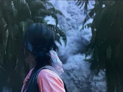 The image displays the upper back and right profile of an individual, positioned facing away from the viewer and looking towards the left. The person has dark hair styled in a low ponytail or bun, secured with a light-colored, possibly pink and white patterned scrunchie or fabric tie. A light blue or turquoise bandana or headscarf is visible wrapped around the head, partially obscured by the hair. The individual wears a light-colored, likely pink or peach, long-sleeved shirt, layered underneath a darker, possibly black or dark blue, sleeveless garment resembling a vest. A small, circular earring is visible on the right earlobe.
The background consists of dense, dark green foliage, appearing to be large-leafed plants or palm trees, framing a lighter, blue-grey expanse. This background exhibits indistinct, voluminous forms consistent with dense cloud cover, mist, or disturbed water. The overall lighting of the scene is subdued. The specified location is the city of Apshak, Uzbekistan.