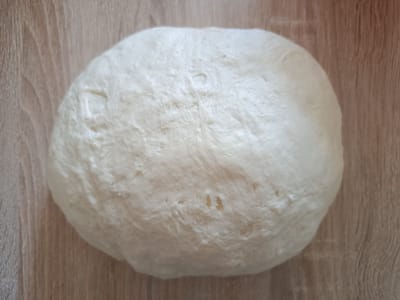 A ball of raw dough rests on a light brown wooden surface, likely a countertop. The dough is pale white and appears to be slightly risen, with visible air pockets and uneven texture on its surface. The wooden surface exhibits a grain pattern with variations in light and dark brown hues. The overall scene is simple and focused on the dough. This image was captured in Градска општина Палилула, Serbia.