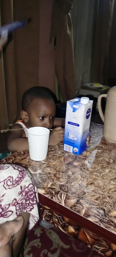 In a dimly lit indoor setting, likely a home in Rijiyar Zaki, Nigeria, a young Black child with short hair and dark eyes is seated at a patterned table. The child is wearing an orange and white tank top and appears to be looking down and to their left, possibly engaged with something on the table. In front of the child is a white cup and a carton of milk with blue and white branding. To the right of the milk carton, a beige-colored thermos is partially visible. The table surface has a mottled brown and gold pattern. In the background, brown curtains hang, and a hint of a shelf with a plate and yellow object is discernible. To the far left of the frame, a blue and black object, possibly a pen or marker, is held by an unseen hand, pointing towards the brown curtains. The overall scene suggests a casual domestic moment.