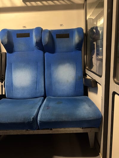 Two blue fabric train seats are visible in an interior setting, likely a train car. The seats are upholstered and appear worn, with some lighter patches indicating wear or sun fading. Each seat has a headrest with a dark rectangular patch on it, possibly for a label or identification. The seats are arranged side-by-side. A portion of a window is visible to the right, reflecting light and an indistinct silhouette of a person. The background wall is a neutral color. There is a small rectangular dark object on the right seat cushion. Numbers "78" and "71" are visible on the wall above the seats, possibly indicating seat numbers. The scene is well-lit, with the light source appearing to be from above. The image does not contain any elements that specifically place it in the city of Dalgopol, Bulgaria.