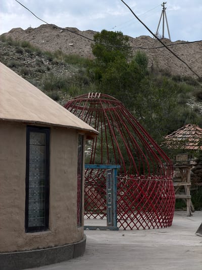 In the village of Кызыл-Туу, Kyrgyzstan, a partially constructed red yurt stands on a paved surface. The yurt's wooden lattice structure is visible, as is its framework of thin red poles. To the left, the curved wall of a completed structure, possibly another dwelling, is seen. This structure has a tan, rough-textured exterior and a dark-framed window with patterned curtains. In the background, a steep, rocky hillside is covered with sparse vegetation. A power line stretches across the frame, and a utility pole is visible on the crest of the hill. Another small structure with a tiled roof is situated to the right of the red yurt. The sky is overcast.