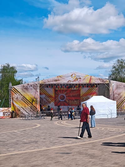 The image shows a public square in Uglich, Russia. A stage has been erected, decorated with banners celebrating the 9th of May, Victory Day in Russia. In front of the stage are several individuals, including two wearing military uniforms with red berets. An older man, wearing a red shirt and dark pants, walks across the square using a cane. A white tent stands near the stage. The sky is blue with scattered clouds.
