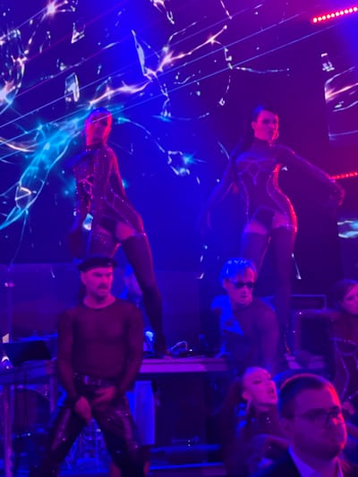 This is a photograph taken indoors, possibly at an event or performance venue. The lighting is predominantly blue and pink, suggesting stage lighting. In the background, a large digital screen displays a fractured, abstract image with bright blue and white lines and shapes.

In the foreground, there are several people. Two women in the upper half of the frame are wearing sparkling, dark-colored outfits that appear to be bodysuits or leotards with attached stockings. Both have their hair styled and appear to be performing or posing. The woman on the left has her head tilted back slightly, and the woman on the right is looking forward with her mouth open.

Below the two women, there are several individuals in the lower half of the frame. On the left, a man wearing a black beret and a sheer black long-sleeved shirt with black pants is visible. He appears to be looking towards the left of the frame. Next to him, another person with dark hair and sunglasses is looking to the right. Further to the right, a woman with her hair in a ponytail is looking upwards. In the bottom right corner, a man wearing glasses and a dark jacket is looking towards the right.

Various objects are present, including what appear to be musical instruments or equipment, such as keyboards and a mixing board, situated on a table-like structure in the middle ground. The overall scene suggests a lively performance or entertainment setting. The image is noted as being taken in Tsilkani, Georgia.