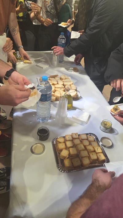 A table is covered with a white tablecloth. Several people stand around the table. Various items on the table include two bottles of water, plastic cups, two open jars, a tray of small toasted bread cubes, a plate of larger toasted bread pieces, a bottle of olive oil, and napkins. People appear to be making and eating small toasts, possibly with toppings from the jars. The location is likely Altura, Spain, given the prompt context, though specific details in the image do not confirm this.