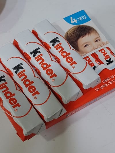 Four individually wrapped Kinder chocolate bars are presented within their original retail packaging on a plain, light-colored surface. The white cardboard portion of the packaging features a close-up image of a child's face, partially obscured by a Kinder chocolate bar. The "Kinder" logo is prominently displayed in red on both the individual bar wrappers and the outer packaging. Korean text, including "4개입" (4 pieces), "내용량: 50g" (net weight: 50g), and "1회 섭취참고량 (15g)" (1 serving reference amount: 15g), is visible on the packaging. The four bars are arranged side-by-side, resting in a red, U-shaped tray that forms the base of the retail package. The items are stationary. This scene is located in 서대문구, South Korea.