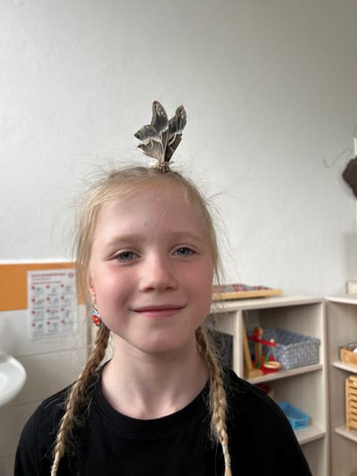 A young girl with blonde hair in two braids and blue eyes is looking at the camera with a slight smile. A large moth is perched on top of her head, its wings spread open. She is wearing a black t-shirt and a colorful earring on her left ear. The background is a brightly lit room with white walls and shelving units containing various items, including baskets, toys, and what appears to be an educational chart on the left wall. The overall impression is a playful and candid moment captured indoors. The context of Zlín, Czechia, is not explicitly visible in the image itself.