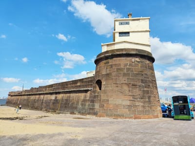 The image depicts a seaside scene in Wallasey, United Kingdom. In the foreground, a sandy beach meets a paved area. Dominating the mid-ground is a large, curved stone wall, likely part of a historic fortification or sea defense, constructed from rough-hewn, reddish-brown stones. Atop the curved section of the wall, a multi-story, cream-colored building stands, featuring dark rectangular windows and a small chimney. Several seagulls perch on the building's roof and ledges.

To the right of the stone structure, a bright blue trailer, possibly a food vendor or mobile unit, is parked, with a person visible near its opening. A blue car is parked behind the trailer, and a Union Jack flag flies on a pole in the distance. In the far left of the image, two people are walking on the sandy beach, one wearing a green top and a patterned skirt, the other in a white top.

The sky is a vibrant blue, dotted with fluffy white clouds. Two small white streaks, likely contrails from aircraft, are visible in the upper left quadrant, and several seagulls are flying in the sky. The scene suggests a bright, sunny day, likely mid-day, given the shadows. The overall mood is relaxed and leisurely, indicative of a pleasant day out by the coast. No visible text is present in the image, other than what might be on the blue trailer which is too small to read.