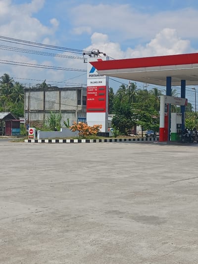 The image displays a Pertamina gas station located in Keudealuerheung, Indonesia, under a clear blue sky with scattered white cumulus clouds. A large concrete forecourt dominates the lower half of the scene.

A tall white pylon sign, centrally positioned, bears the "PERTAMINA" logo at its apex. Below the logo, the number "14.242.104" is displayed, followed by three digital panels indicating fuel types: "PERTAMAX TURBO 98," "PERTAMAX 92," and "DEXLITE."

The gas station structure consists of a prominent red and white canopy supported by blue and white pillars, covering multiple fuel dispensers. Several motorcycles are parked near the rightmost pumps. A blue sedan is partially visible in the mid-ground, behind the pumps.

To the left of the pylon sign, a landscaped median contains small bushes with distinctive orange flowers and various green plants, bordered by a black and white striped curb. A red circular prohibitory sign with a white horizontal bar is present on the median, alongside a blue sign with "BSI Bank Aceh" visible text.

The background features dense tropical foliage, including numerous palm trees. Behind the left side of the gas station, a multi-story, unfinished or dilapidated concrete building with exposed rebar is visible. Overhead power lines traverse the upper portion of the sky. No individuals are prominently engaged in specific actions within the frame.