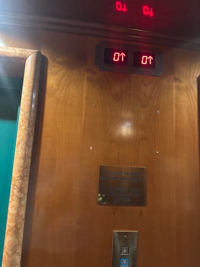 The image shows an elevator panel with illuminated numbers and arrows. Two red digital displays above the panel show "01" with downward-pointing arrows. Below these, a rectangular panel with two illuminated "01" displays and upward-pointing arrows is visible. A metallic plaque with text in Italian and English is mounted on the wooden panel, reading "In caso d'incendio non usare gli ascensori" and "In event of fire please do not use the lifts". A single elevator button panel with up and down arrow buttons is located below the plaque. The elevator panel is set within a wooden wall with a marble-effect trim on the left side. This scene is located in San Martino Buon Albergo, Italy.
