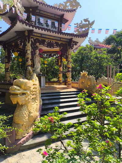 The image depicts a Buddhist temple in Tay Nha Trang Ward, Vietnam. The foreground features a golden dragon statue and lush green foliage with pink flowers, leading to a set of stone stairs. Beyond the stairs is an ornate pagoda with intricate carvings and dragon motifs, adorned with colorful flags. A statue of a woman stands within the pagoda, surrounded by yellow and pink decorative spheres. The temple complex is set amidst a dense green environment, with more structures and trees visible in the background. The sky is clear and blue.