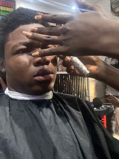 A person is lying supine with a black barber cape on, with a white barber paper collar around their neck. A hand holds a small metal container above the person's face. This appears to be a barber applying product to the person's face. The location is possibly Ogwashi-Uku, Nigeria, as per the prompt's request.