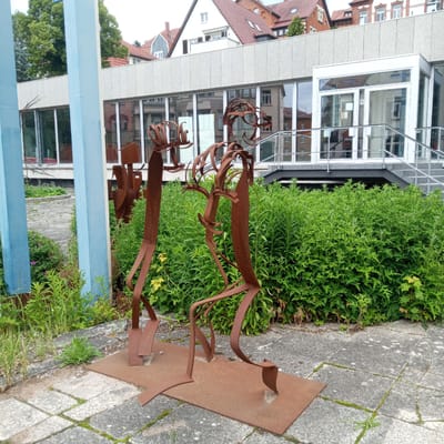 In Eisenach, Germany, on a paved area with scattered moss, two abstract metal sculptures stand. The sculptures, made of rusted metal, are human-like figures with thin, bent limbs and abstract heads. The figure on the left appears to be holding a shovel-like object. Both figures are placed on a rectangular metal base. Behind the sculptures is a dense green hedge, and beyond that, a modern building with large windows reflecting the surrounding environment. The building has a light-colored, textured facade and a glass entrance with a staircase leading up to it. In the background, several traditional German houses with red tiled roofs are visible, and trees with green foliage. The sky is overcast.