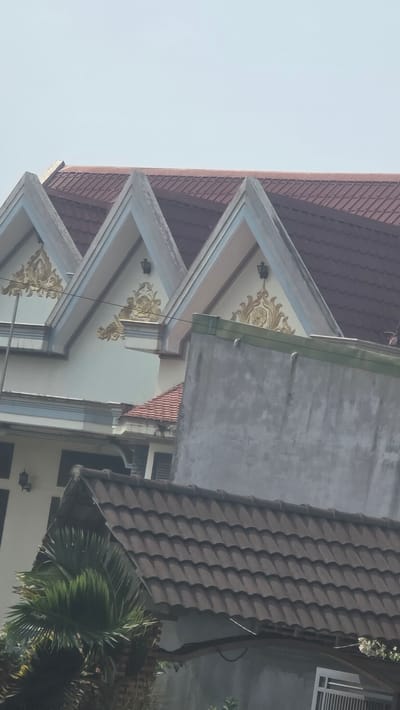 The image presents a close-up, angled view of building architecture, possibly in the city of Trung Hồ, Vietnam. It features layered roof designs with ornamental golden patterns adorning the eaves. One roof segment is covered in dark brown tiles, while another structure displays a gray concrete façade. The sky is visible in the upper left corner, suggesting an outdoor setting. A partially visible tree is situated towards the bottom right of the frame.