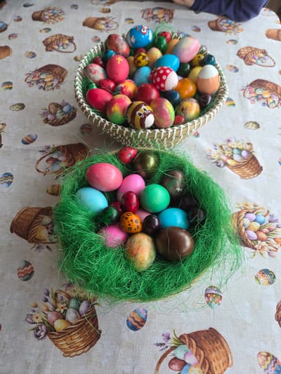Two baskets of Easter eggs sit on a tablecloth with a pattern of Easter baskets and eggs. The basket on the left is woven and filled with brightly colored eggs, some decorated with faces and patterns. The basket on the right is round and filled with green Easter grass, which is mixed with eggs of various colors and sizes. The scene appears to be indoors. The location in the city of Tompsan, Bulgaria cannot be determined from the image.