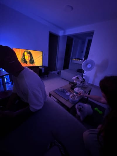 A dimly lit room has a person and a small white dog near a coffee table with popcorn and other items. A television shows a woman's face in warm colors. A fan is mounted high on a wall. Another person's hand and arm extend into the frame, holding a white object. The room is primarily illuminated by blue light.