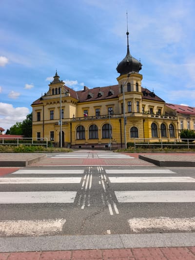 The image shows a pedestrian crossing in front of a yellow building with a dark-colored turret in Vysoké Mýto, Czechia. The building has multiple windows and a red tile roof. A flag, possibly the Czech flag, is visible on a balcony on the building. The sky is blue with scattered clouds. The foreground features the white stripes of the pedestrian crossing on an asphalt road. Red bricks are visible along the edges of the road and sidewalk. Trees and a white fence are present behind the crossing. No people or vehicles are visible in the image. The overall scene depicts a public urban space on a clear day.