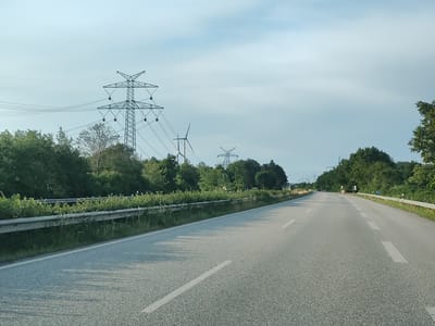 The image depicts a view of a paved road in Damlos, Germany, under a cloudy sky. The road appears to be a multi-lane highway or a major road, with white lane markings visible. To the left of the road, there is a metal guardrail separating it from a grassy area with various green bushes and trees. In the background, several tall electricity pylons with wires stretching between them are prominent. One of the pylons is particularly large and intricate. Further in the distance, a wind turbine is visible amongst the trees, suggesting a rural or semi-rural landscape. The trees form a dense line along the roadside and in the background, with some taller trees interspersed. The overall scene suggests a bright, possibly overcast day. There are no visible people or vehicles actively engaged in any interactions in the foreground.