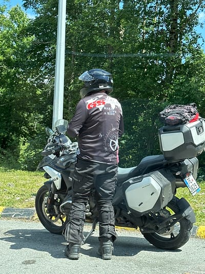 A person wearing a black motorcycle helmet and a black long-sleeved shirt with red and white graphics stands next to a black BMW GS motorcycle. The shirt has the words "MAKE LIFE A RIDE" printed on it, along with a compass graphic. The person is wearing black motorcycle pants and black boots. The motorcycle has two silver panniers and a black top case with a red and black bag strapped to it. A license plate, "7E 3629," is visible on the rear of the motorcycle. The scene appears to be on the side of a road, with trees and greenery in the background. The lighting suggests it is daytime, with sunlight casting shadows. The image was captured in Crkvine, Montenegro.