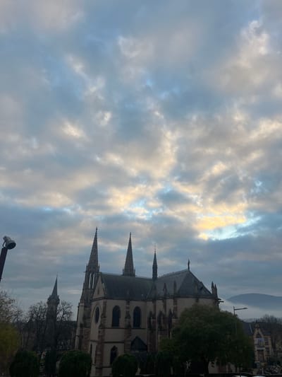 This image depicts the Église Saints-Pierre-et-Paul (Church of Saints Peter and Paul) in the city of Obernai, France, under an overcast sky. The prominent church structure occupies the midground and right-center of the frame, featuring light-colored stone facades, dark gray slate roofs, and three tall, pointed spires with intricate detailing. A smaller, darker pointed spire, likely belonging to another historic building such as the Kappelturm, is visible further in the background to the left.

The sky is filled with varying shades of gray clouds, with patches of lighter yellowish-orange and faint blue visible, indicating diffused sunlight. In the foreground, to the left, a street light fixture with a dark pole and a rectangular lamp is partially visible. Several trees with sparse, dark foliage are present around the church and in the foreground, alongside manicured shrubs. To the right of the church, additional buildings with dark roofs and light walls are partially visible, and a distant mountain range, partially obscured by low-lying fog or mist, forms the horizon. No individuals or discernible actions are present in the scene.