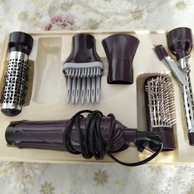 The image displays a beige plastic tray containing a set of hair styling tools. The tools are primarily a deep purple color with silver accents. From left to right, the objects include a round brush attachment with bristles and ventilation holes, a wide comb attachment with many narrow teeth, a hair dryer handle with a coiled black cord plugged into it, a smaller cylindrical brush attachment with stiff bristles, and what appears to be a curling iron attachment. The setting is a flat surface covered with a light-colored fabric featuring a floral pattern in white and pale yellow. The lighting suggests an indoor environment, possibly a home, as there are no indications of outdoor conditions. The overall arrangement suggests the items are new or neatly stored, possibly in their original packaging or a storage case. There is no visible text in the image.