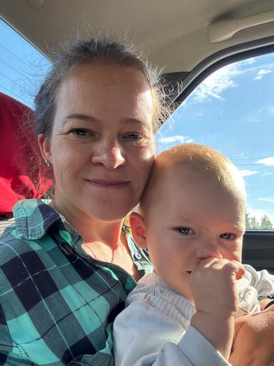 A woman and a baby are in the front seat of a car, with the woman taking a selfie. The woman has graying hair pulled back and is wearing a teal and navy plaid shirt. The baby, who has blond hair and blue eyes, is looking at the camera with its hand in its mouth. The background shows a blue sky with white clouds and some trees. The image appears to have been taken on a sunny day. The car's interior, including the headliner and window trim, is visible. The overall setting is a daytime outdoor scene, possibly in the city of Zarnitsa SNT, Russia, given the context.