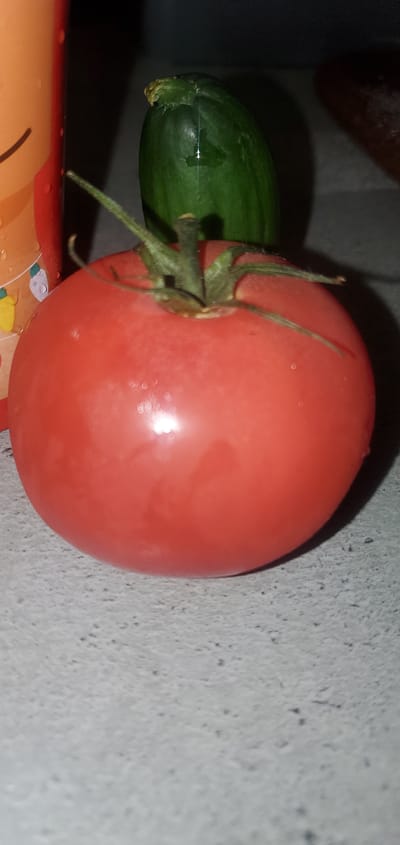 The image shows a close-up of a ripe, red tomato with water droplets on its surface, resting on a speckled gray countertop. To the left, a portion of an orange container with a smiley face design is visible, also with water droplets. Behind the tomato, a green cucumber is partially visible, its skin exhibiting vertical ridges. The lighting suggests an indoor setting, casting shadows on the countertop. No specific location in Sasbach, Germany can be determined from the visual information alone.