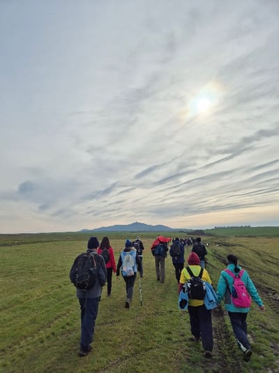 A group of approximately 15 to 20 individuals is observed hiking across an open, green grassy field near Stara Reka, Bulgaria. The sky is largely overcast with horizontal cloud formations, and the sun appears as a bright, partially obscured disc with a faint halo in the upper right quadrant of the frame. The hikers, mostly adults, are seen from behind, walking in a loose formation along a wide, unpaved dirt and grass track that traverses the field. They are dressed in various outdoor and casual clothing, including jackets, pants, and hats. Most individuals carry backpacks of different colors, notably black, blue, pink, red, and yellow. One person in the middle ground utilizes a walking stick. In the far distance, a range of hills or mountains is visible against the horizon. On the peak of the most prominent distant hill, a distinct, large circular concrete structure, consistent with the Buzludzha Monument, is clearly identifiable. The terrain appears slightly uneven, with some darker patches indicating possibly wet or muddy ground.