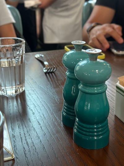 The image depicts an indoor dining setting. The foreground features a dark brown wooden table with visible grain. On the table, from left to right: a clear glass tumbler, approximately half-filled with clear liquid; a three-tined silver-colored metal fork resting face down; and two matching teal-colored spice grinders. The grinder on the right has a silver finial embossed with the letter 'P' and "LE CREUSET" embossed on its base, while the grinder on the left has a silver finial embossed with an 'S'. To the far right, a portion of a white rectangular holder with green stripes, containing beige paper packets, is visible. In the blurred background, partial figures of at least two individuals are present. One individual on the left is wearing a white short-sleeved garment. Another individual on the right is wearing a dark short-sleeved garment, displaying a tan arm with a dark bracelet, and is shown interacting with a smartphone that has a visible yellow protective case. The lighting appears to be ambient.