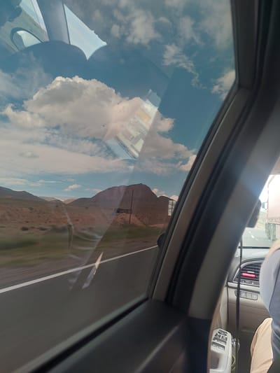 The image, taken from within a vehicle looking out a side window, captures a journey through an arid, mountainous landscape under a bright, sunny sky.

In the foreground, the dark frame of the car window partially encloses the view. To the right, sections of the car's interior are visible, including part of the dashboard, air vents (one prominently displaying a small red object, possibly an air freshener or indicator light), and a panel with various buttons. A white remote control device with multiple buttons rests on the passenger side. A person's torso and legs, dressed in a light top and beige bottoms, are partially visible in the upper right, suggesting they are a passenger or driver. The window itself features prominent reflections, including large, white, fluffy clouds and the silhouette of another person's head and shoulders.

Beyond the window, a paved road with a distinct white line stretches into the distance. It is bordered by dry, reddish-brown mountains or hills that dominate the middle ground. Faint utility poles and wires can be discerned against the distant mountainous peaks. The sky above is a clear blue, adorned with scattered white clouds, some of which are strongly reflected in the window.

The scene conveys the activity of traveling by car during daylight hours on a clear, bright day. The strong reflections are a notable detail, creating a layered visual effect where the interior and exterior environments merge. The arid terrain and mountainous features are consistent with the geography of Armenia, particularly the region around Paruyr Sevak. No discernible text is visible in the image.