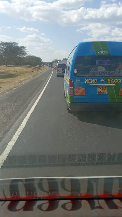 A line of vehicles, mostly minibuses, is stopped on a two-lane road. The minibus closest is blue and green with the words "IMANI SHUTTLE" and "NENO SACCO" printed on the back. The number plate reads "KDL 460S". The road is surrounded by dry vegetation and scattered trees. The sky is bright with scattered clouds, and the image is taken from inside another vehicle, with part of the dashboard visible at the bottom.